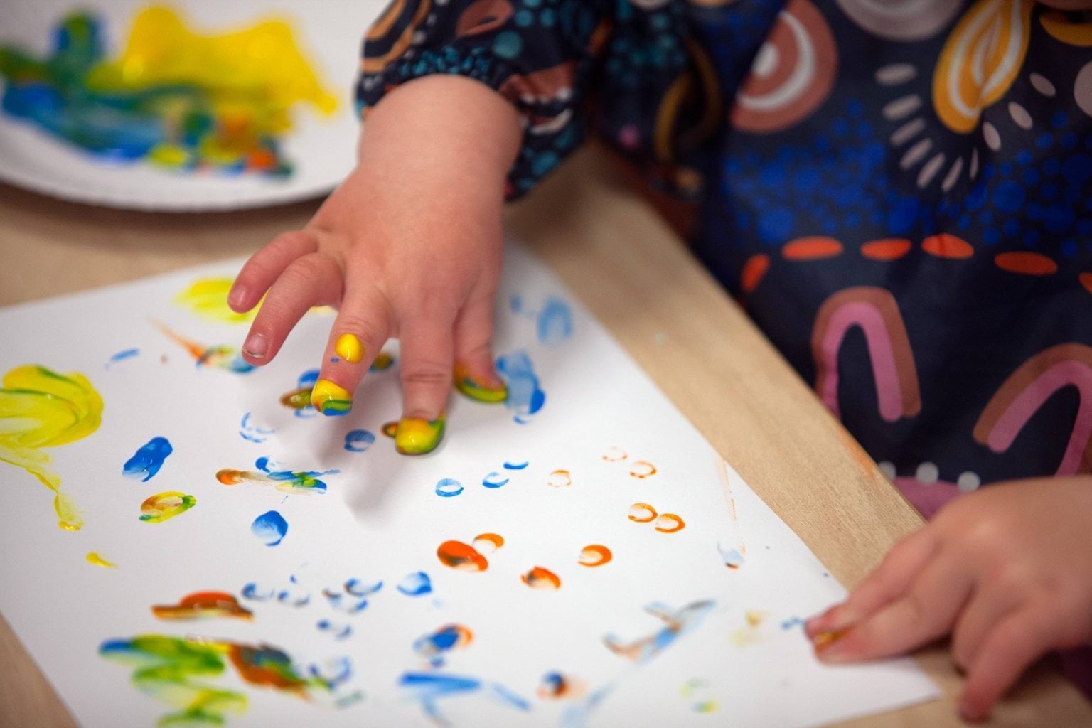 Close-up of a child’s hands finger painting at a wooden table at Early Learning Victoria Bani Walup (Murtoa), wearing a smock with colourful Aboriginal art design.