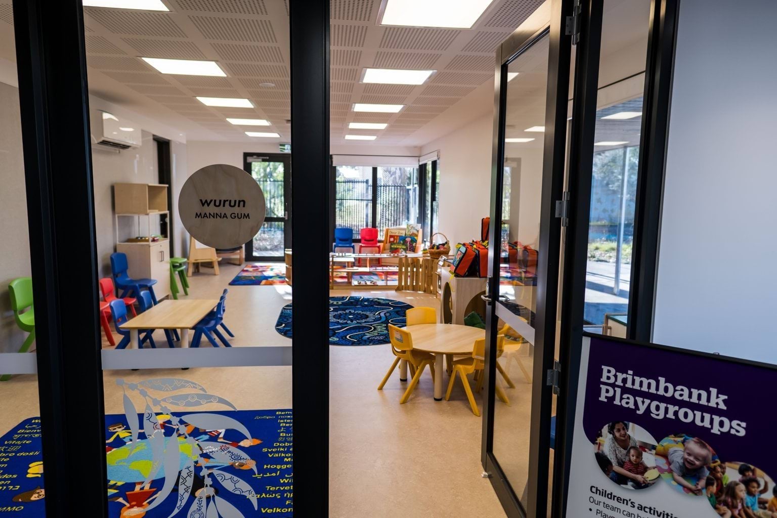 View through glass doors, one open, of the wurun (manna gum) room at Early Learning Victoria Muyan. Bright blue multicultural and Aboriginal art rugs dot the floor, along with small wooden tables with colourful chairs. 