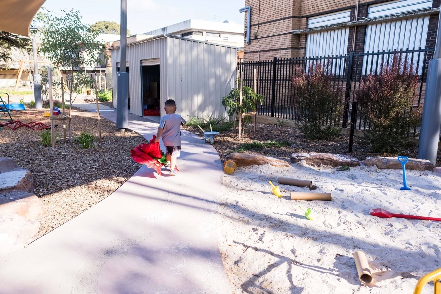 View of outdoor area at Early Learning Victoria Muyan, with sandpit and toys, and a young boy running along a path towards the tanbark play area. 