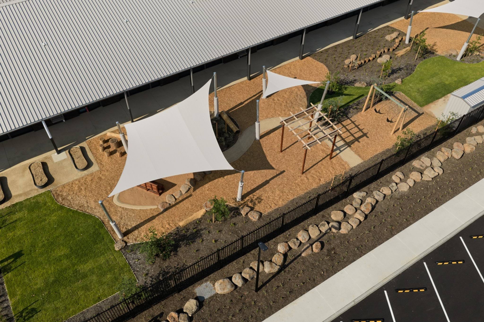 An aerial view of an early learning outdoor play area with shade sails, climbing equipment, sand play zones, and surrounding garden beds.